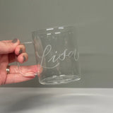 A hand holding a clear glass mug with the name 'Lisa' engraved on it.