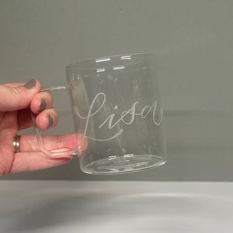 A hand holding a clear glass mug with the name 'Lisa' engraved on it.