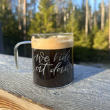Kaffemugg i glas, graverad med texten We ride at Dawn. Hangraverad, står på ett trädäck med sol och skog i bakgrunden. 