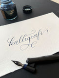 Bild på ordet kalligrafi skrivet i blått bläck på vitt papper med en traditionell kalligrafipenna