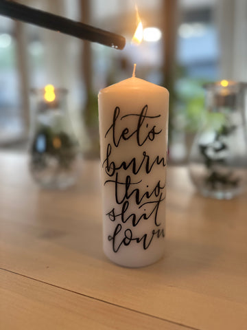 A white candle with text on its side that reads 'Let's burn this shit down', placed on a wooden surface with a black holder and a lit wick.