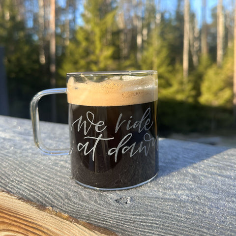 A clear glass mug with hand-engraved text 'We ride at dawn' on it