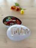 Handmålat vitt påskägg i plåt, fullt med påskgodis. Locket är handmålat med namnet Tindra i guld. 