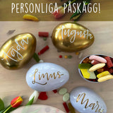 Guldfärgade och vita påskägg i plåt, handtextade med namn som gör dem personliga. Godis och tulpaner i bakgrunden. 