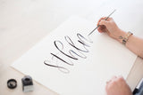 Bild på hand som skriver Sthlm i kalligrafi med pensel och svart bläck, workshop i Lettering i Stockholm
