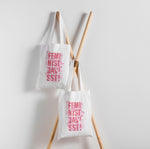 Två tygkassar som hänger mot en vit vägg, men rosa text och illustrationer. Feminist Javisst!