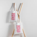 Två tygkassar som hänger mot en vit vägg, men rosa text och illustrationer. Feminist Javisst!