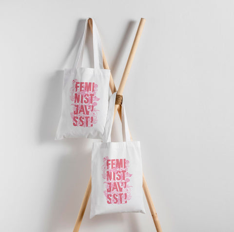 Två tygkassar som hänger mot en vit vägg, men rosa text och illustrationer. Feminist Javisst!