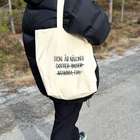 En person som bär en väska som det står "hon är någons dotter, syster, mamma, fru" på, men där allt utom "hon är någon" är överstruket.