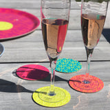 A set of four colorful glass coasters with summer-themed patterns, placed under two glasses filled with a pink beverage, on a wooden table.