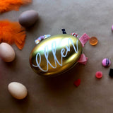 Ett guldfärgat påskägg av plåt med Ellen handskrivet i vit oljefärg på locket. Oranga fjädrar och godis i bakgrunden