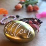 Ett guldfärgat påskägg i plåt med texten Ellen på locket. Fullt med påskgodis och tulpaner i bakgrunden. Namn skrivet i vit kalligrafi 